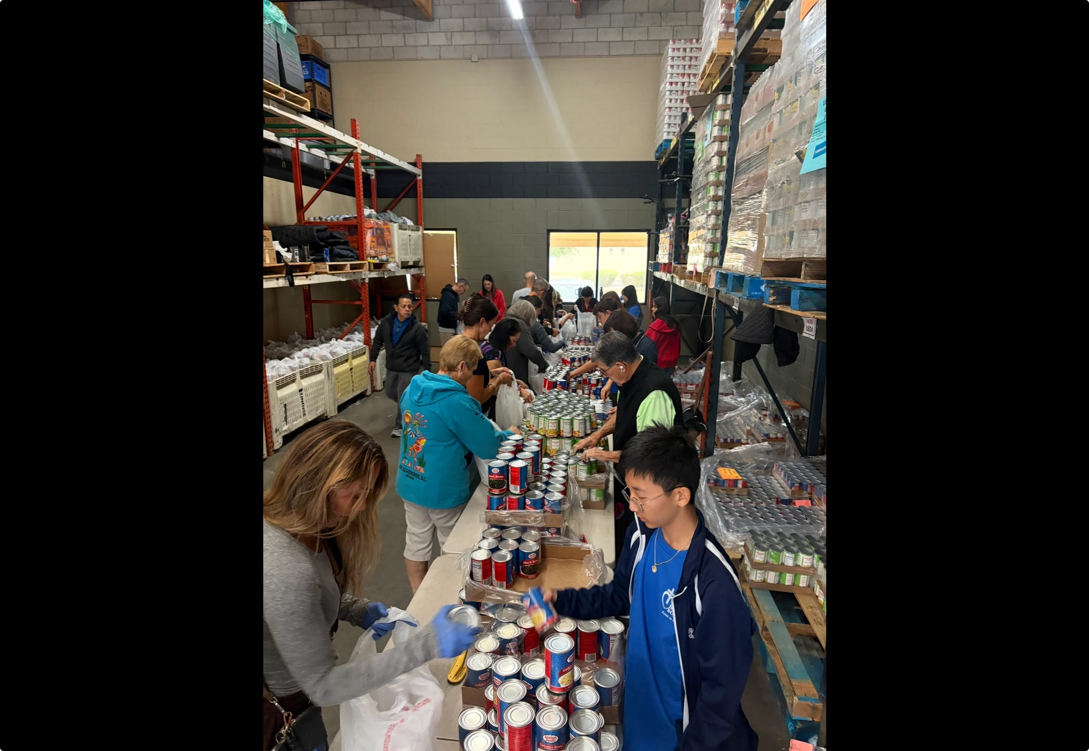 A diverse group of volunteers lines up along a table in a warehouse, actively sorting and packing hundreds of canned goods into plastic bags for distribution.